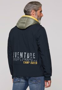 Marinejacke mit einer grün-gelben Kapuze, die auf der Rückseite weißen und gelben Text aufweist. Glatte Textur und maßgeschneiderte Passform.