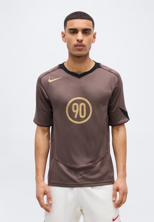 Νέος άνδρας που φοράει καφέ αθλητική μπλούζα Nike με χρυσό αριθμό "90" στο μπροστινό μέρος και λευκό σορτς, σε στάση μπροστά σε απλό φωτεινό φόντο.