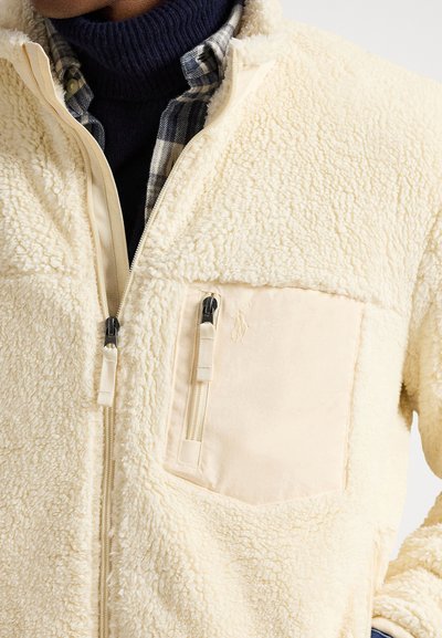 Κοντινό πλάνο ενός ατόμου που φοράει εκρού σακάκι από φλις με φερμουάρ στην τσέπη στο στήθος, πάνω από μπλε πουκάμισο καρό και μπλε ζιβάγκο.