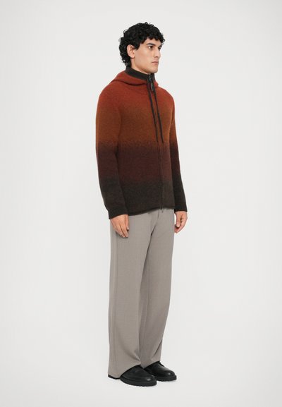 Sweater com capuz e fecho éclair em tons degradê de laranja a castanho, feito de material suave. Combinado com calças largas cinza claro e sapatos pretos.