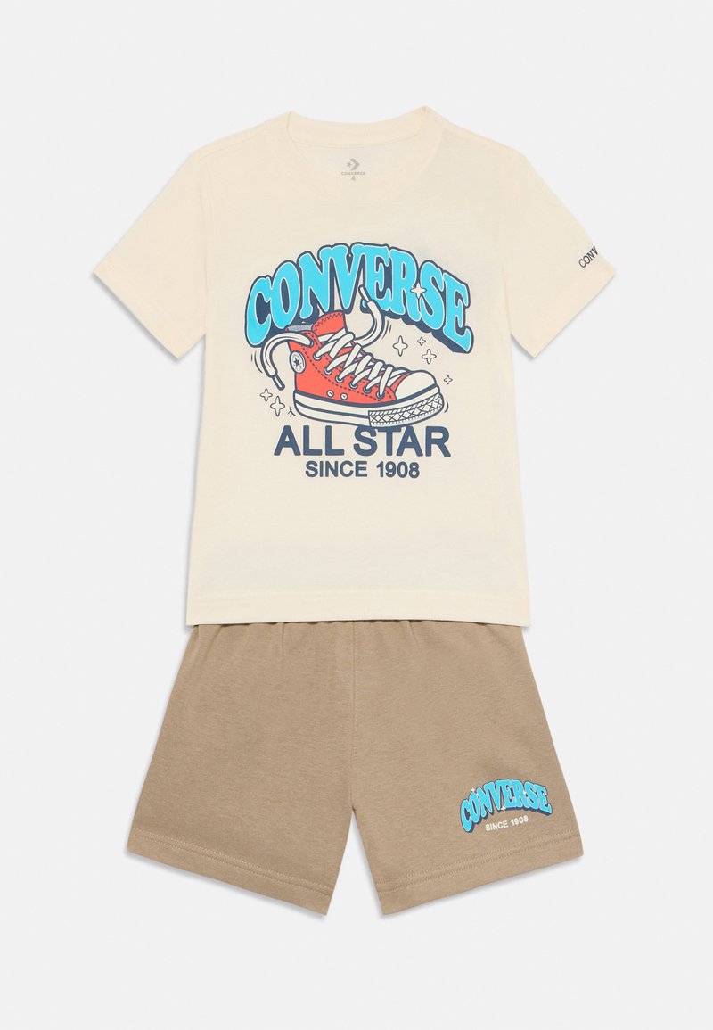 Krems krāsas īsām piedurknēm krekls ar zilu un sarkanu sporta apavu grafiku un tekstu "CONVERSE ALL STAR SINCE 1908". Komplektu papildina bēša šorti.
