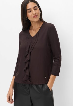 Vrouw met een donker langemouwen top met geplooide voorkant en een zwarte broek, staand tegen een effen witte achtergrond.