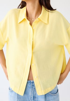 Frau trägt ein lockeres, gelbes Kurzarmhemd mit Knöpfen über hellblauen Jeans, mit einer goldenen Halskette, die am Hals sichtbar ist.