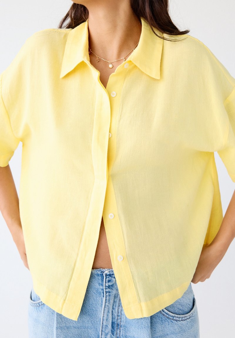 Femme portant une chemise jaune ample à manches courtes boutonnée sur un jean bleu clair avec un collier en or visible au niveau du cou.