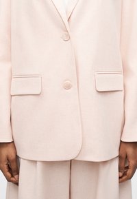 Ρουμπινί ροζ σακάκι ραμμένο, με δύο κουμπιά και τσέπες με καπάκι, φορεμένο πάνω από αντίστοιχο παντελόνι με φαρδιά πόδια, με τα χέρια χαλαρά στα πλάγια.