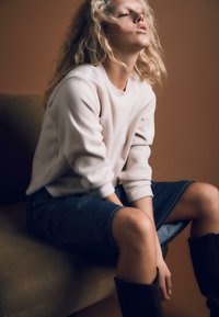 Jasno beżowy sweter z dzianiny z prążkowanym dekoltem, zestawiony ze spódnicą midi z denimu. Modelka siedzi, prezentując teksturowany materiał i luźny krój.