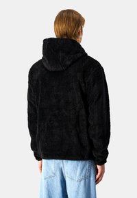 Chaqueta con capucha de forro polar negra con una superficie suave y texturizada. Presenta un corte relajado y una capucha grande, combinada con pantalones de mezclilla azul claro.