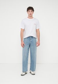 Λευκό t-shirt με κοντά μανίκια και κόκκινη εκτύπωση, συνδυασμένο με ανοιχτό μπλε τζιν παντελόνι φαρδιάς γραμμής. Τα παπούτσια έχουν μαύρες και λευκές λεπτομέρειες.