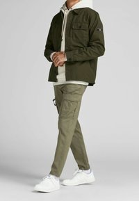 Olivgrön jacka med knappade fickor, lager på lager över en ljusgrå huvtröja. Matchande olivgröna cargo-byxor och vita sneakers fulländar klädseln.