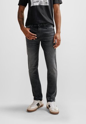 Mężczyzna w wyblakłych czarnych jeansach slim fit, białych i beżowych trampkach oraz czarnym koszulce z nadrukiem, stojący na jednolitym tle.