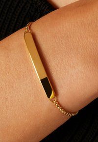 Gouden armband met een platte rechthoekige staaf, voorzien van een gepolijste oppervlakte en een gestructureerde ketting. De staaf heeft aan één uiteinde een zwart accent.