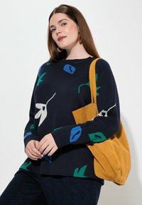 Donna con lunghi capelli castani che indossa un maglione blu navy con motivi floreali, pantaloni scuri, orecchini intrecciati e porta una borsa a tracolla in suede giallo senape.