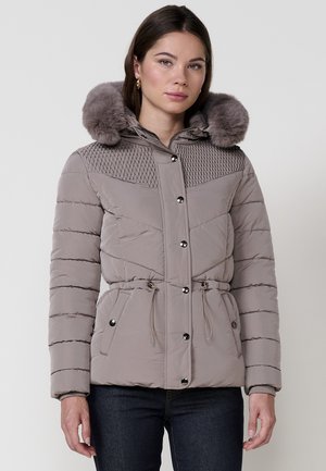 Mujer con chaqueta acolchada color topo y capucha con borde de pelo, botones a presión, cintura con cordón y bolsillos laterales, de pie frente a un fondo simple.