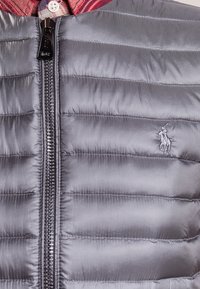 Šedá péřová bunda s hladkou, lesklou texturou, která má vodorovně prošívané sekce a černý zip. Vyšitý logo na hrudi.