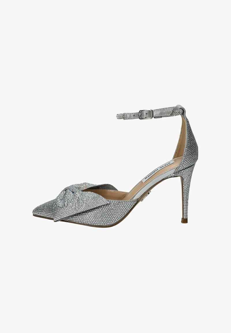 Zapato de tacón alto plateado con punta afilada, adornado con brillantes, una correa en el tobillo con hebilla y un detalle anudado en la parte delantera.