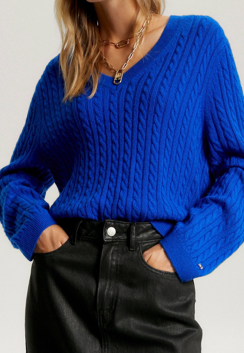 Pull bleu en maille torsadée avec col en V, doté de poignets côtelés, associé à un pantalon noir taille haute, souligné par un bouton métallique visible.
