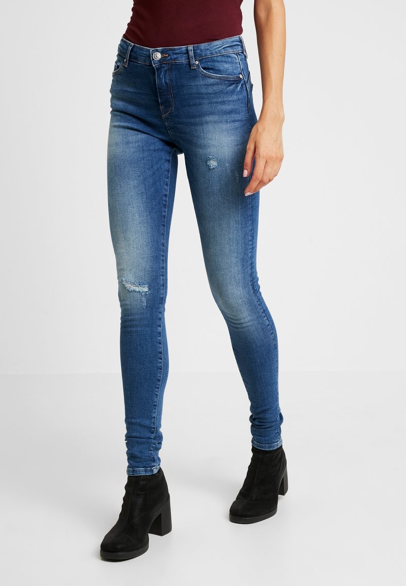 Femme portant un jean skinny bleu délavé avec une légère usure et des bottines à talons noires, debout devant un fond uni.
