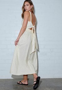 Vestido de color beige claro con lunares negros, espalda abierta y un lazo atado. Diseño fluidos y largo hasta el tobillo con una textura suave.
