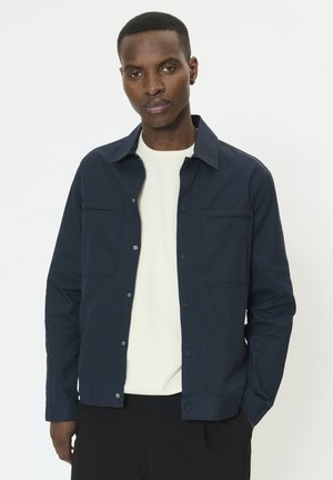 Man draagt een marineblauwe jas met knopen over een wit overhemd, staand met één hand in de zak tegen een effen witte achtergrond.