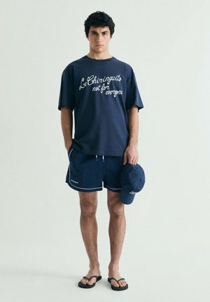 Joven con camiseta azul marino con texto blanco, pantalones cortos a juego, chanclas negras, sosteniendo una gorra azul marino, de pie frente a un fondo liso.