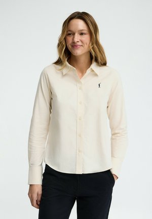 Donna che indossa una camicia color crema con bottoni e un piccolo logo scuro di un giocatore di polo sul petto, e pantaloni neri, con le mani rilassate ai lati.
