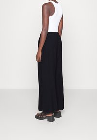 Pantalons noirs à jambe large en tissu texturé, avec une taille élastique, associés à des sandales noires à lanières. Modèle présenté de dos.