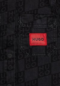 Camicia in denim nera con motivo testurizzato del logo "HUGO". Presenta una tasca sul petto con etichetta rossa e bottoni neri. Design del colletto classico.