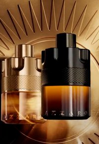 Deux flacons de parfum Azzaro, un doré et un noir avec un liquide ambré, sur un fond radial métallique doré.
