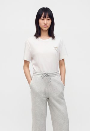 Joven mujer con cabello liso y oscuro, que lleva una camiseta blanca y pantalones deportivos gris claro con las manos en los bolsillos, sobre un fondo liso.