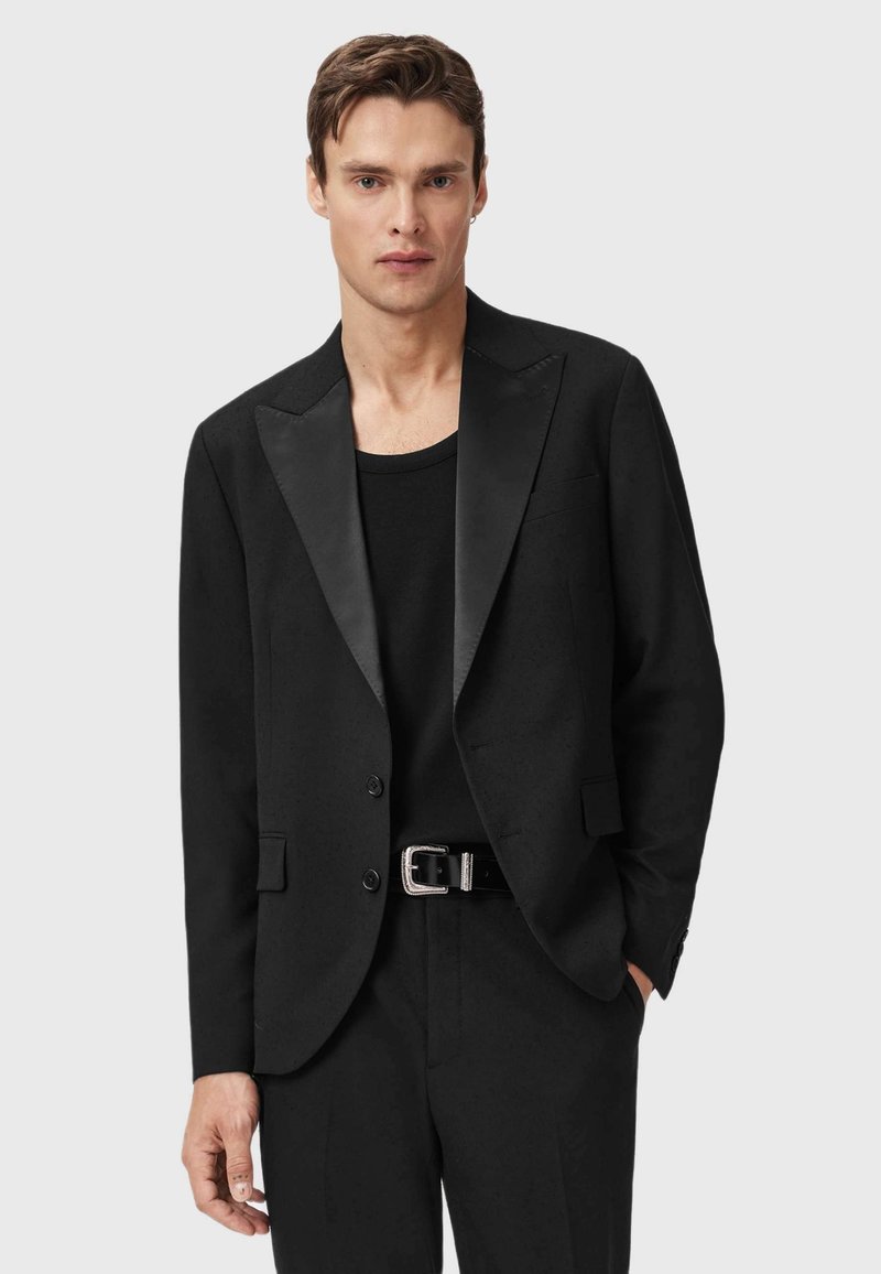 Uomo che indossa una giacca nera con rever in satin, camicia nera, pantaloni neri e cintura nera con fibbia argento, in posa su uno sfondo grigio.