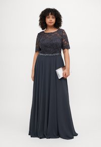 Rochie de seară lungă, albastră marină, cu un corsaj din dantelă, mâneci scurte și detalii cu mărgele la talie. Fustă fluidă cu o textură de chiffon. Accesoriu tip clutch argintiu.