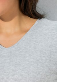 Maglietta grigia chiara con scollo a V, realizzata in tessuto morbido, con un logo a forma di cuore ricamato in modo discreto sull'area sinistra del petto.