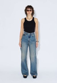 Musta hihaton toppi, korkeavyötäröiset vaaleansiniset suorat farkut ja mustat kengät. Mallilla on ruskeat aurinkolasit, lyhyet kiharat hiukset ja hopeiset korut.