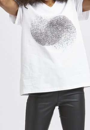 Person trägt ein weißes T-Shirt mit V-Ausschnitt und schwarzem abstraktem Kreis-Muster sowie schwarze Kunstlederhosen.
