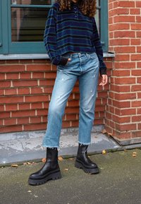 Polo de manga larga a rayas en azul marino y verde, vaqueros ligeros de mezclilla desgastados con dobladillos deshilachados y botas de tobillo de plataforma negras.