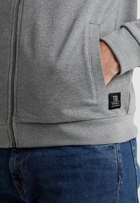 Mano nella tasca di una felpa grigia con zip, con etichetta nera "TTO Original Clothing", abbinata a jeans blu.