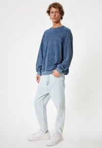 Sudadera de cuello redondo azul claro con tela texturizada, combinada con unos vaqueros de corte ancho de lavado claro y zapatillas blancas. Detalles sutilmente desgastados en los vaqueros.