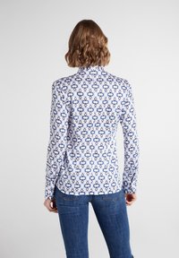 Bílá košile s dlouhými rukávy a modrým geometrickým vzorem, má límeček a manžety na knoflíky, doplněná modrými džínami.