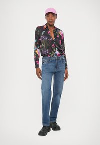 Camicia nera a fiori con colori vivaci, abbinata a jeans blu e scarpe nere. Il modello indossa un berretto rosa chiaro.