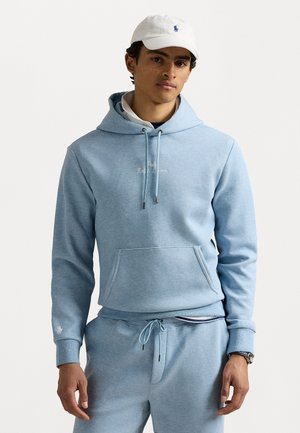 Hoodie bleu clair en tissu doux avec capuche à cordon, poche avant et logo brodé. Porté avec un pantalon de survêtement assorti et une casquette blanche.