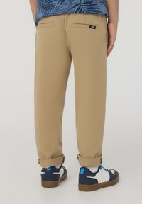 Chinos color khaki con taglio dritto, dotati di due tasche posteriori, risvolti e indossati con sneakers navy e bianche. Etichetta del marchio visibile sulla vita.