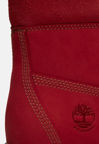 Rode suède laarssectie met drievoudige tan stiknaden en een geperst boomlogo met het label "waterdicht" op het zijpaneel.