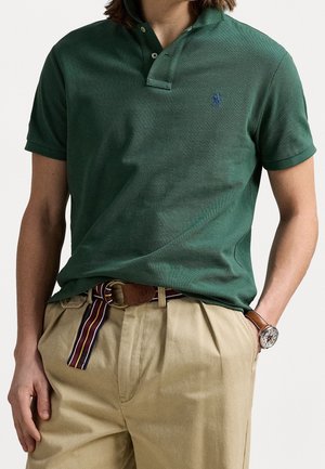 Homme portant un polo vert foncé à manches courtes et un pantalon plissé beige avec une ceinture rayée et une montre-bracelet, une main dans la poche.
