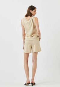 Beige ärmelloses Oberteil mit drapiertem Rückendetail, kombiniert mit passenden Shorts. Das Model trägt schwarze Sandalen mit offenem Zeh. Glatter Stoff, lässiges Design.
