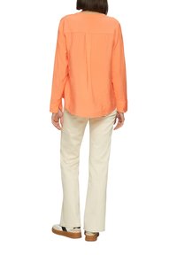 Chemise orange à manches longues avec des plis au dos, associée à un pantalon évasé blanc cassé et des baskets décontractées avec des accents noirs.