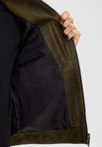 Olijfgroene suede jas aan de binnenkant, met een gladde zwarte voering en een zijzak. Rits sluiting langs de rand, met een gestructureerde stof.