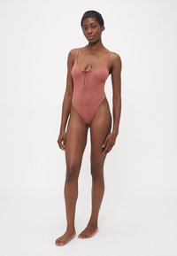 Woman standing barefoot wearing a sleeveless, dusty rose one-piece swimsuit with a front tie detail against a plain white background.