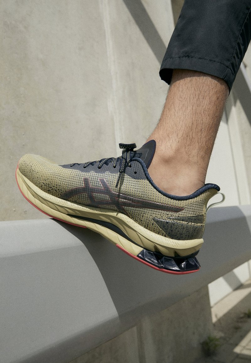 Chaussure de course avec une tige en mesh beige respirant, des accents bleu foncé, et une semelle texturée avec des touches rouges et un design arrière unique.