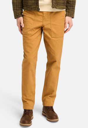 Brown cotton pants with large front pockets, straight-leg design, and visible stitching. Worn with brown lace-up boots.
