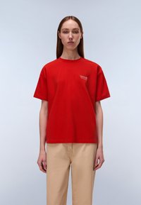 Κόκκινο βαμβακερό μπλουζάκι με κοντά μανίκια, στρογγυλή λαιμόκοψη και μικρή λεπτομέρεια λογότυπου στο αριστερό στήθος. Συνδυάζεται με μπεζ παντελόνι, εμπρός όψη.